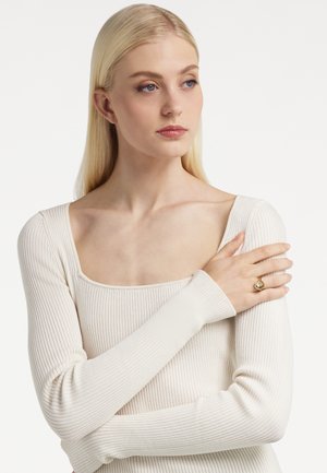 Plavuša u kremasto rebrastoj majici dugih rukava sa kvadratnim izrezom, lijeva ruka joj je naslonjena na desno rame, nosi zlatni prsten.