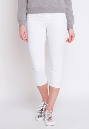 Personne portant un pantalon blanc court, un haut gris à manches longues et des baskets métalliques argentées, debout devant un fond uni.