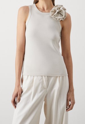 Helle beige gerippte Tanktop mit einem strukturierten Stoff, rundem Halsausschnitt und einem großen Stoffblumenakzent auf der linken Schulter. Kombiniert mit lockeren Hosen.