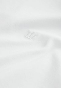 Valge kangas, mille keskel on peenelt tikitud krooni logo, näidates pehmeid kortse ja siledat tekstuuri.