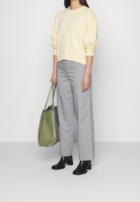 Sweatshirt jaune clair avec marquage blanc, pantalon large gris et bottes à talons carrés noires. Sac fourre-tout vert avec des bordures bleues.