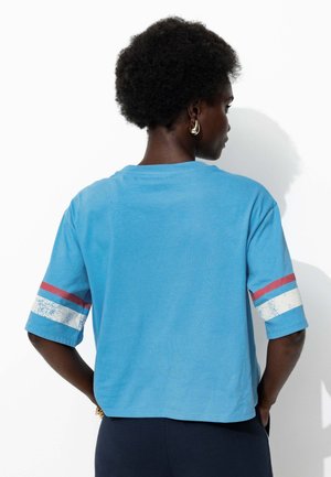 Une personne avec des cheveux bouclés courts porte une chemise bleue avec des rayures rouges et blanches sur les manches, se tenant avec les mains dans les poches de son pantalon sombre, tournant le dos.