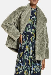 Chaqueta de piel sintética de color verde oliva, esponjosa, con un amplio y suave cuello y hombros caídos, combinada con un vestido estampado que presenta colores azul marino, verde y azul claro.