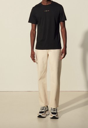Czarny t-shirt z krótkim rękawem z kolorowym logo, beżowe spodnie o prostym kroju i czarno-białe trampki z kolorowymi akcentami.
