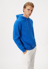 Blå huvtröja gjord av mjukt material, med en framficka, justerbara snoddar och ribbade ärmslut. Bärs med ljusa byxor.