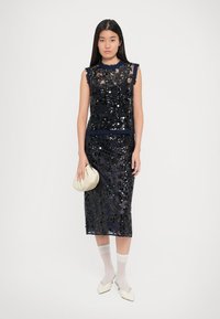 Donna che indossa un vestito senza maniche nero con paillettes, tiene in mano una piccola borsa beige, calze bianche e scarpe a punta color crema, in piedi contro uno sfondo semplice.
