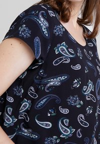 Top de tela negra con mangas cortas, que presenta un patrón de paisley en tonos azules y verdes, con acentos florales y un escote en forma de barco.
