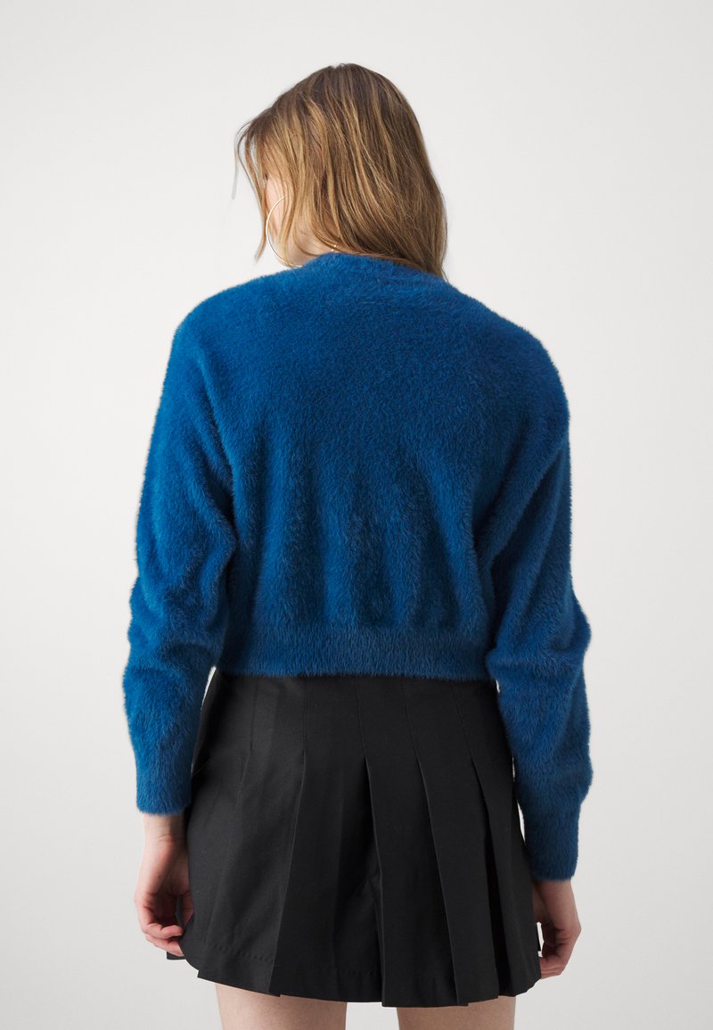 Pullover blu soffice con maniche lunghe, tessuto strutturato, orlo corto, abbinato a una gonna nera plissettata, che mostra una silhouette semplice e moderna.