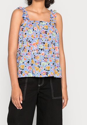 Vrouw die een blauwe, mouwloze top met bloemenprint en gerimpelde bandjes draagt, gecombineerd met een zwarte broek met witte stiknaden.