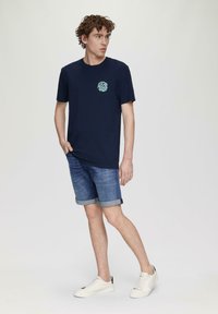 Marineblauwe katoenen T-shirt met een ronde mintgroene logo, gecombineerd met spijkerbroekshorts en witte sneakers. Casual pasvorm en ontspannen stijl.