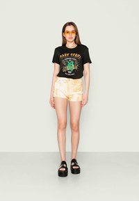 T-shirt gráfica preta com impressão multicolorida, acompanhada de calções de pêssego tie-dye. Sandálias de plataforma pretas completam o conjunto.