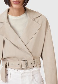 Beige Wildlederjacke mit breiten Revers, tailliert und strukturiert, getragen über einem hellen Oberteil und weißen Hosen.