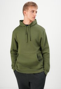 Young man looking right, wearing an olive green hoodie with front pocket and dark gray pants, standing against a white background.