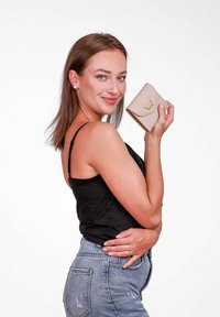 Světle béžová peněženka se zlatým logem, hladkým povrchem a zakřiveným designem klopy, držená v ruce osoby na neutrálním pozadí.