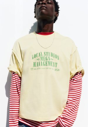 Giovane persona di colore che indossa una maglietta color crema con scritte verdi sopra una lunga camicia a righe rosse e bianche, posando contro uno sfondo bianco.
