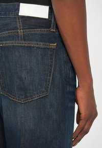 Jeans de mezclilla azul oscuro con costuras naranjas, que presentan un bolsillo trasero y una etiqueta blanca en la cinturilla. Textura suave y corte clásico.