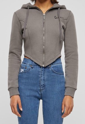 Vrouw draagt een strakke grijze cropped zip-up hoodie met trekkoordjes en een hoge taille blauwe spijkerbroek tegen een effen achtergrond.