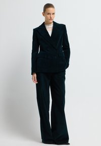 Tmavě zelený kordový oblek s dvouřadým blejzrem a širokými kalhotami. Hladká textura, přizpůsobený střih a detaily na předních kapsách.