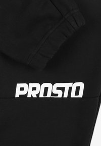Czarne spodnie dresowe o gładkiej fakturze, z wyraźnym białym logo "PROSTO", elastyczną talią i mankietami przy kostkach.