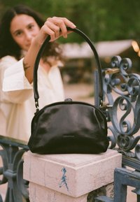 Czarna skórzana torebka o zaokrąglonym kształcie, z krótkim uchwytem i subtelnym detalem w postaci logo. Teksturowana powierzchnia o błyszczącym wykończeniu.