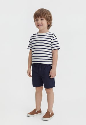 Usmievajúci sa mladý chlapec stojaci, oblečený v bielom tričku s námorníckymi pruhmi, námorníckych krátkych nohaviciach a hnedých nazúvacích topánkach na bielom pozadí.