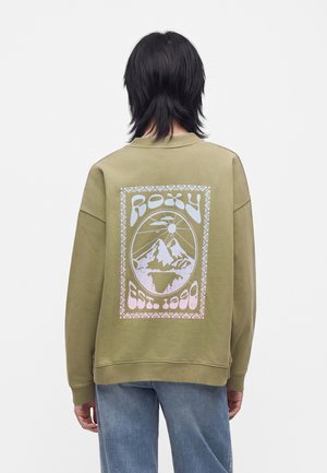 Person med sort hår iført en olivengrøn sweatshirt med en pastelgrafik af bjerge, sol og tekst "ROXY Est. 1990" på bagsiden.