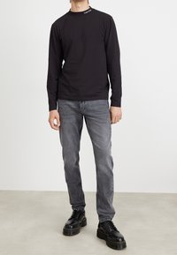 Chemise noire à manches longues avec un col rond et le logo Calvin Klein au niveau du col, associée à un jean droit gris et des chaussures à plateforme noires.