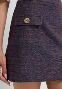Laivasilmuinen tweed-hame punaisilla ja oransseilla langoilla, jossa on etutaskun läppä, jossa on kiiltävä kultainen nappi, pukeutuneena seisovalle henkilölle.