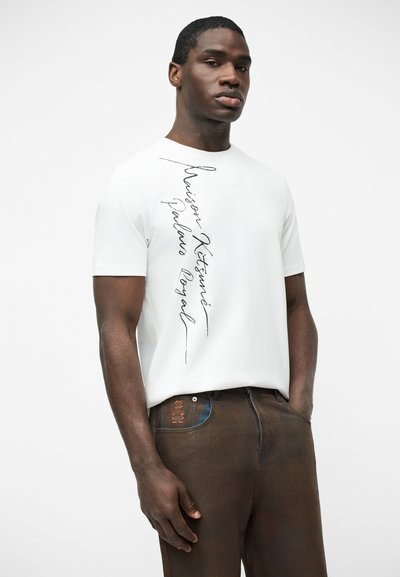 Άντρας με λευκό κοντομάνικο μπλουζάκι με κάθετο κείμενο και καφέ παντελόνι με μικρό κεντημένο σχέδιο κοντά στην τσέπη.