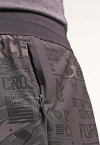 Svarta shorts med en elastisk midja har en diskret grafisk design som inkluderar logotyper och text. Materialet verkar slätt, lätt och flexibelt.