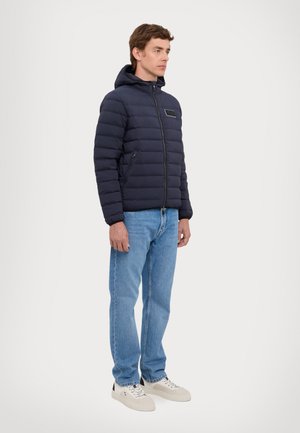 Giubbotto imbottito blu navy con cappuccio, dotato di chiusura zip frontale, tasche laterali e etichetta sul petto. Abbinato a jeans azzurri e sneaker bianche.