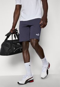 Calções de atletismo azuis com o logótipo branco da Puma, combinados com sapatos brancos com detalhes em vermelho e solas pretas, e uma bolsa de ginásio preta.