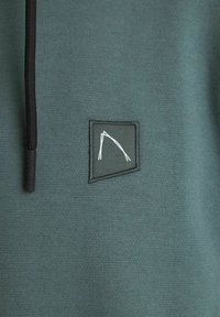 Felpa con cappuccio verde scuro in tessuto texturizzato, con cordini neri e una patch rettangolare con un design astratto bianco cucito nella parte anteriore.