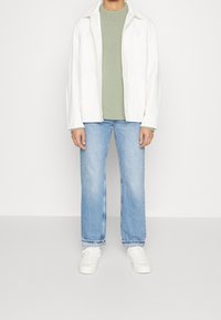 Chemise blanche oversize, pull texturé vert, jean droit bleu et baskets blanches, mettant l'accent sur un design moderne minimaliste.