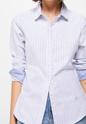Person, die ein weißes und hellblau vertikal gestreiftes Hemd mit hochgekrempelten Ärmeln und blaue Jeans aus Denim trägt.