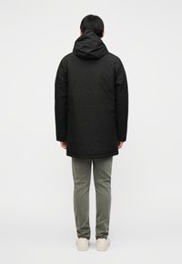 Μαύρο, μονωτικό χειμωνιάτικο παλτό με κουκούλα, με λεία υφή και μήκος που φτάνει μέχρι το μέσο του μηρού. Φορεμένο πάνω από ανοιχτό γκρι παντελόνι.