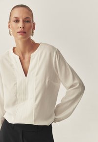 Weiße Bluse aus leichtem Stoff, mit V-Ausschnitt, langen Ärmeln und einem subtilen vertikalen Faltenmuster. Kombiniert mit schwarzen Hosen.