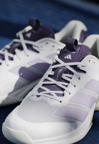 Paire de chaussures de sport blanches et violet clair avec des lacets, des détails en mesh et une languette marquée, reposant sur une surface bleu foncé.