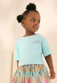 Tricou bleu deschis cu mâneci scurte și imprimat cu „Disney Princess”. Asortat cu o fustă colorată, cu model de prințese, din țesătură transparentă.