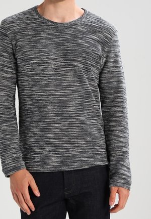 Mies, joka pitää päällään mustavalkoista marled-neulepuseroa pitkine hihoineen ja tummat housut, seisoo yksinkertaisen vaalean taustan edessä.