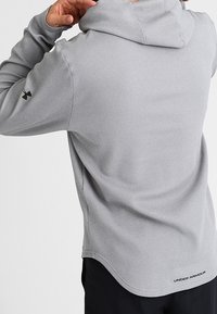 Persona con sudadera de textura gris claro de Under Armour, ajustando la capucha, vista desde atrás con pantalones negros visibles.