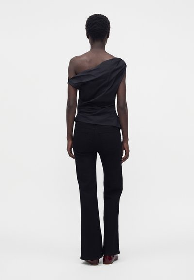 Personne mince portant un top noir une épaule ajusté avec un détail drapé, pantalon bootcut noir et chaussures marron, debout sur un fond blanc uni.