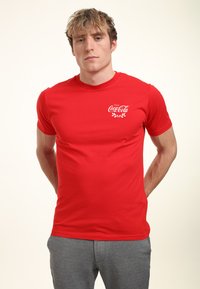 Rød bomulds t-shirt med et hvidt Coca-Cola-logo og kransdesign på venstre bryst. Kortærmet med rund halsudskæring.
