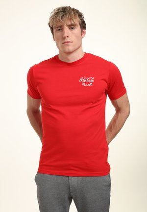 Rode katoenen t-shirt met een wit Coca-Cola logo en een kransontwerp op de linkerkant van de borst. Korte mouwen met een ronde hals.
