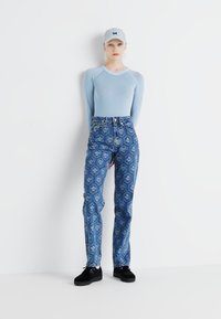 Svetlomodrý top s dlhým rukávom, vzorované modré džínsy s kvetinovými motívmi a čierne topánky. Model pózuje na jednolatom bielom pozadí.