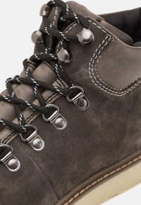 Botín de ante marrón con cordones negros, ojales metálicos plateados, costuras en contraste y suela de goma beige. Primer plano del diseño del zapato.