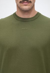 Homme avec une moustache portant un pull en maille vert olive avec le nom de la marque HUGO brodé sur la poitrine.