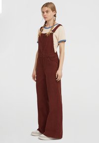 Joven con trenzas rubias que lleva una camiseta beige con ribetes azules y un mono de pana marrón de pernera ancha con cremallera frontal y zapatillas blancas.