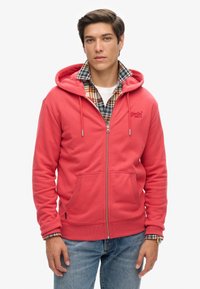 Koraalkleurige zip-up hoodie met een zachte textuur, voorzien van een voorzak, verstelbare trekkoorden en een logo. Gelaagd over een ruitjesoverhemd.