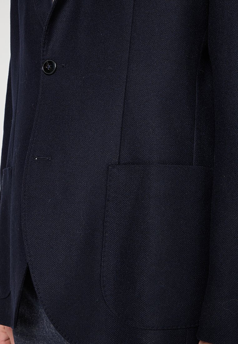 Granatowa wełniana marynarka z pojedynczym zapięciem na guzik, dwiema dużymi kieszeniami z przodu oraz teksturowanym wzorem w jodełkę. Dopasowany krój i elegancki design.
